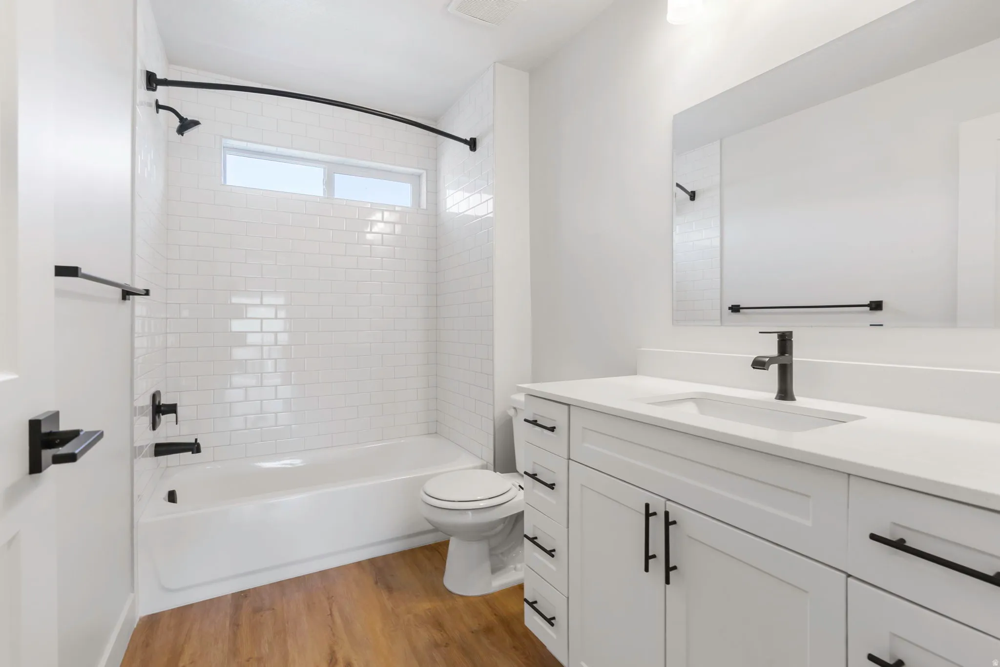 Modern bathroom featuring a single vanity with white cabinetry, a large mirror, and a full bathtub with white subway tile surround and a transom window