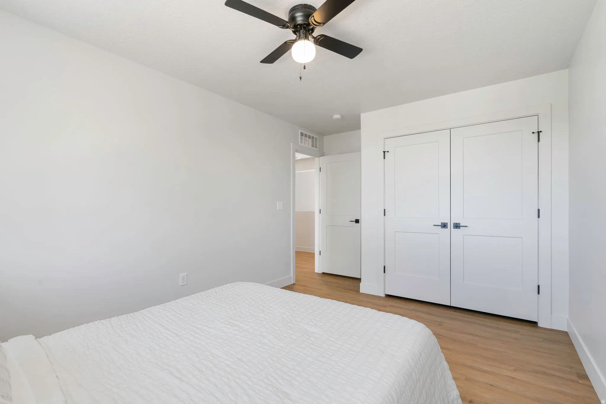 White-painted interior room featuring a ceiling fan with light fixture, light wood-finish flooring, and white double-panel closet doors with dark hardware