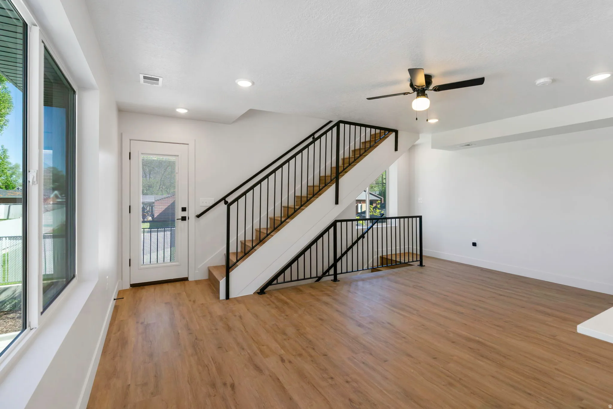 Open-concept living area featuring wood-finish flooring, a contemporary staircase with black metal railings, a full-lite exterior door, recessed lighting, and a ceiling fan