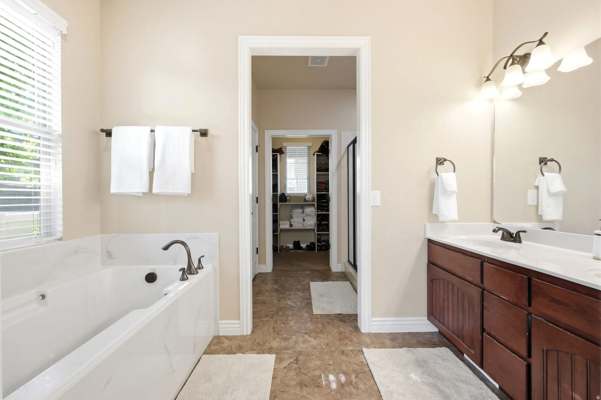 Soaking tub featuring a dark bronze faucet, an adjacent window with blinds, a dark wood vanity with a white countertop and integrated sink, a large mirror, and a multi-bulb light fixture