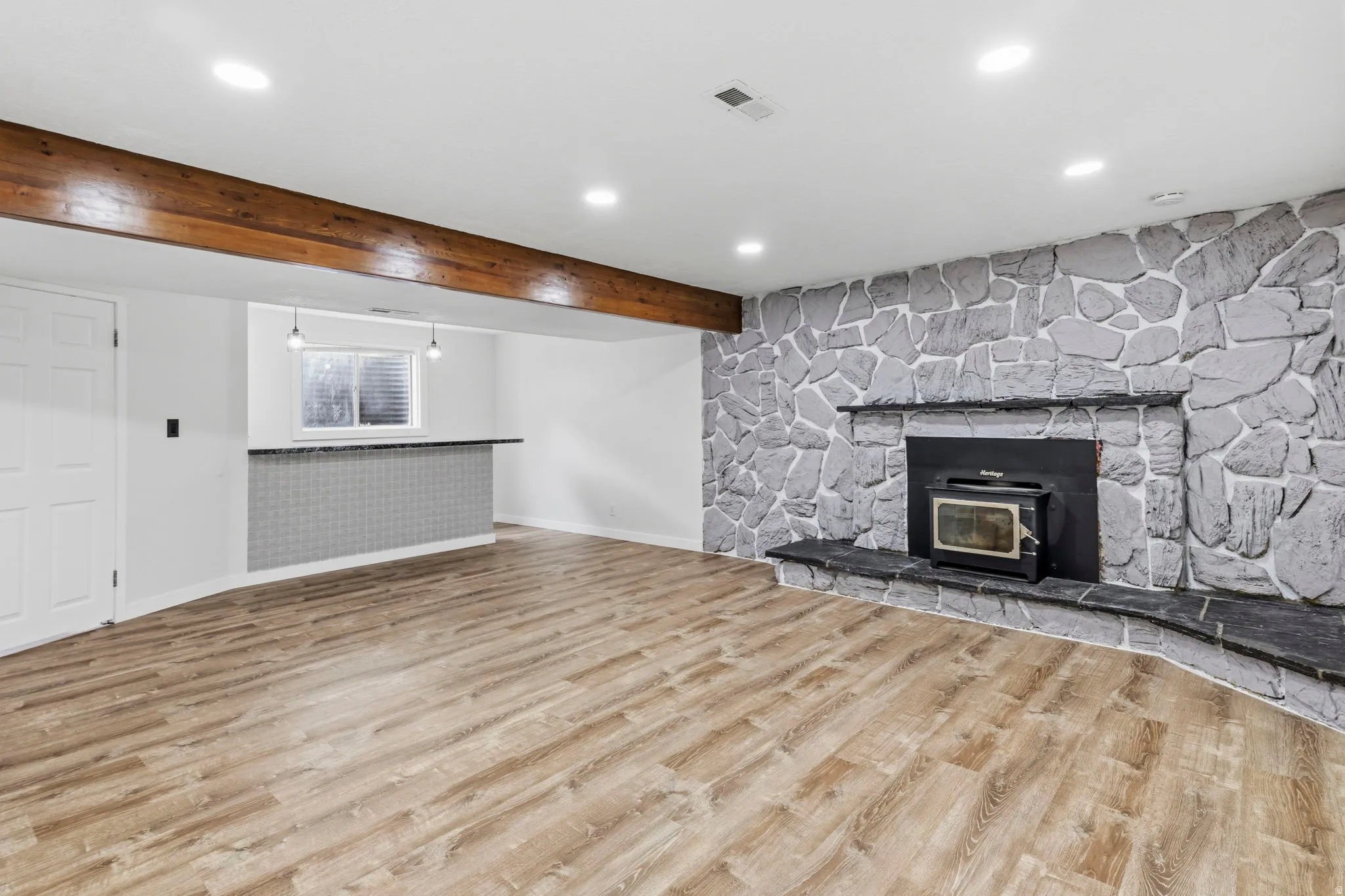 Spacious room featuring wood-finish flooring, a prominent stone fireplace with hearth, a built-in bar with a tile facade and countertop, and exposed wood beam accents