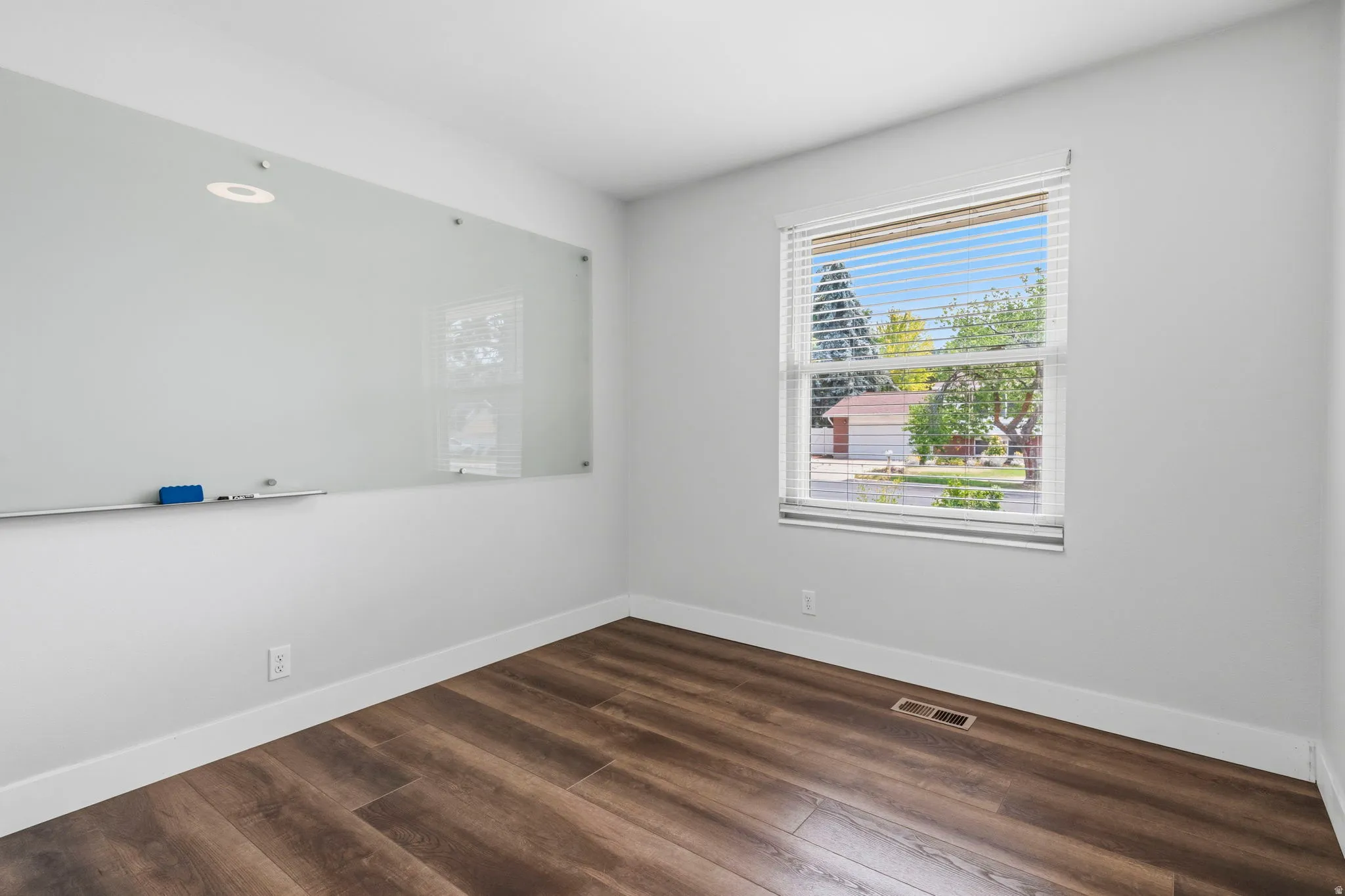 Bright room featuring wood-finish flooring, white baseboards, a large window with blinds, and a wall-mounted glass whiteboard