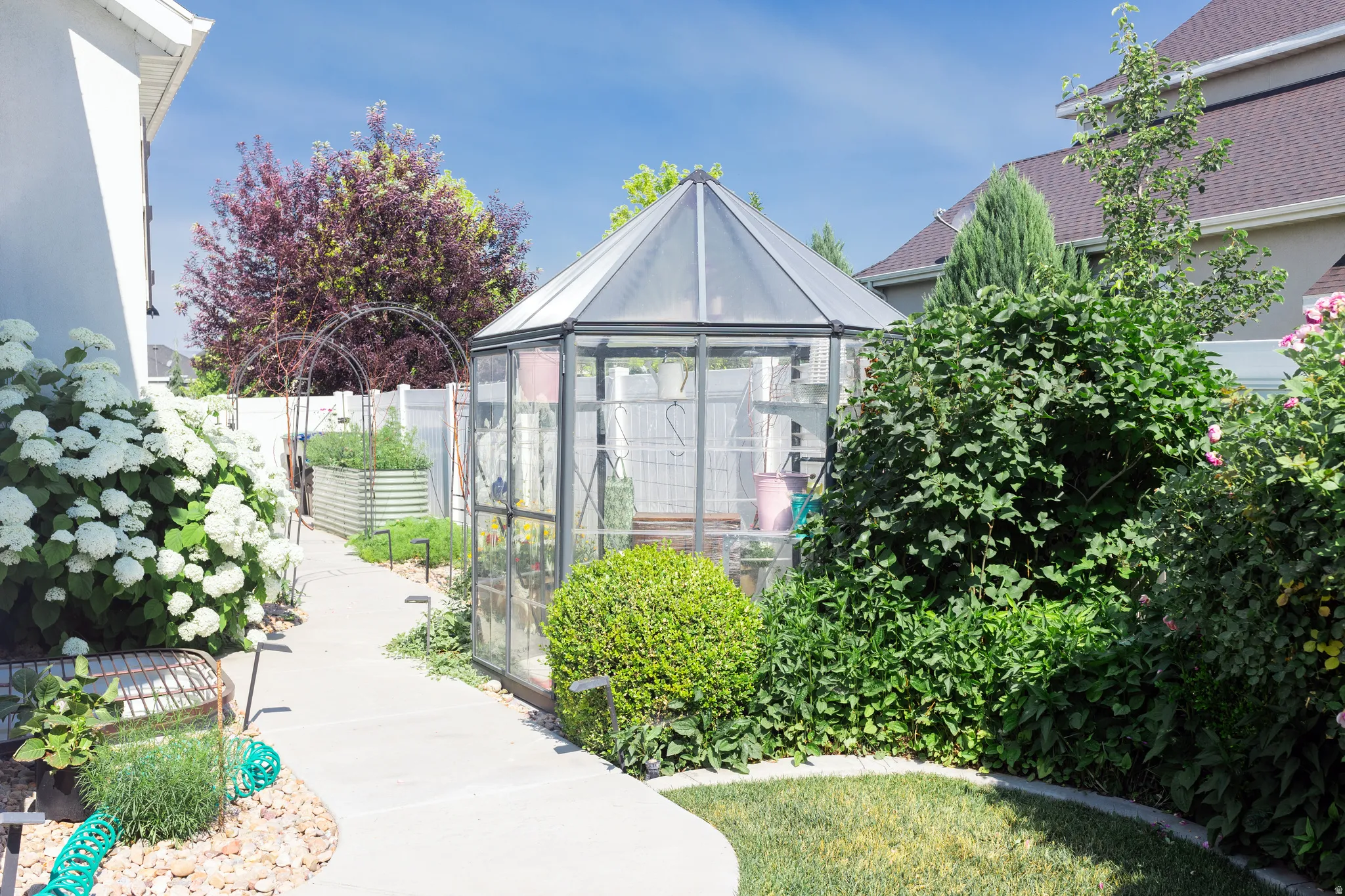 Garden featuring a glass greenhouse, concrete pathway, raised garden beds, and mature landscaping