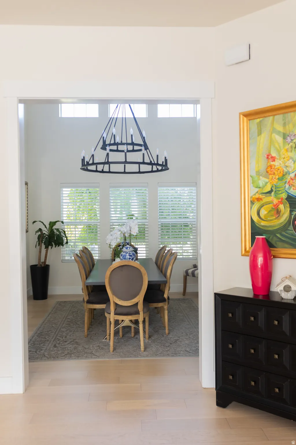 Double-height dining space featuring a large black chandelier, multiple windows with blinds, light wood-finish flooring, and white trim