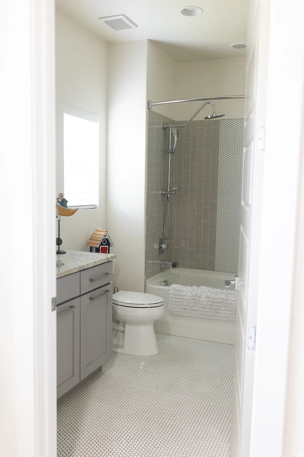 Main floor bathroom featuring white penny tile flooring, a built-in bathtub with subway tile surround, a toilet, and a vanity with a light gray cabinet and solid surface countertop