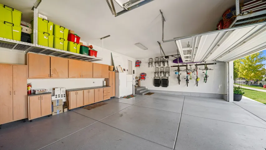 Spacious garage featuring epoxy-coated flooring, extensive wood-finish cabinetry with integrated appliances, overhead storage with bright yellow bins, pendant lighting, and a roll-up garage door opening to natural light and landscaped grounds