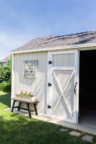 Outdoor storage shed with a shingled roof, light-colored vertical siding, and a white barn-style door with black hardware