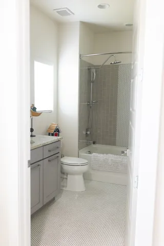 Main floor bathroom featuring white penny tile flooring, a built-in bathtub with subway tile surround, a toilet, and a vanity with a light gray cabinet and solid surface countertop