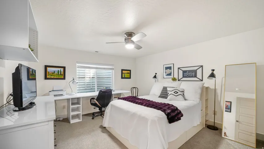 Spacious bedroom featuring a ceiling fan with integrated lighting, generous natural light from shuttered windows, neutral carpeting, and a dedicated workspace with desk and office chair