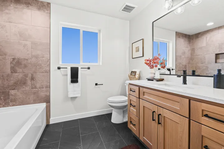 Bathroom featuring a light wood-finish vanity with a white countertop and rectangular sink, black hardware, black floor tiling, and large format wall tiling surrounding the tub