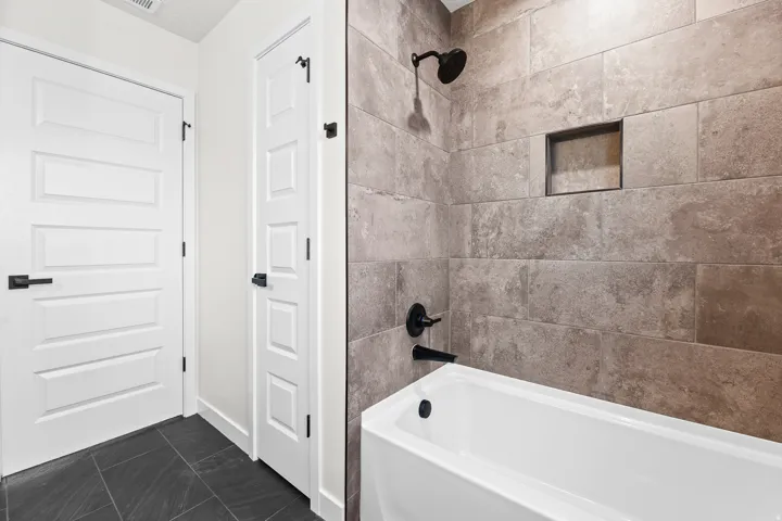 Bathtub with dark-toned tile surround, featuring a built-in shower niche, matte black showerhead, and matching faucet hardware