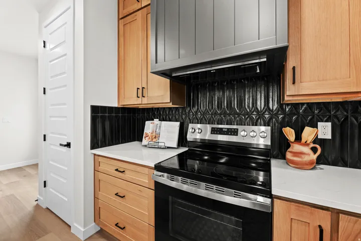 Contemporary kitchen featuring a stainless steel electric range, black decorative tile backsplash, light wood-finish cabinetry, white countertops, and a paneled range hood