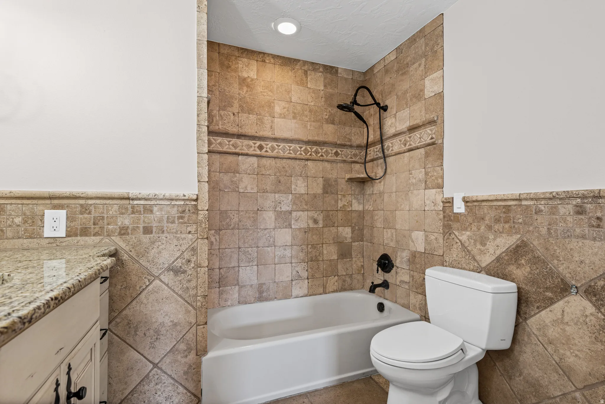 Bathroom featuring a built-in bathtub with a dark-finish shower fixture, tile surround, and a coordinating tiled toilet area