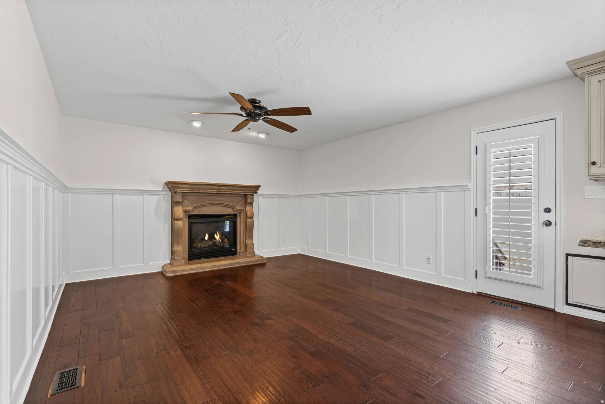 Living space featuring a stone-clad fireplace with a dark insert, dark wood-finish flooring, white wainscoting, and a ceiling fan with integrated lighting