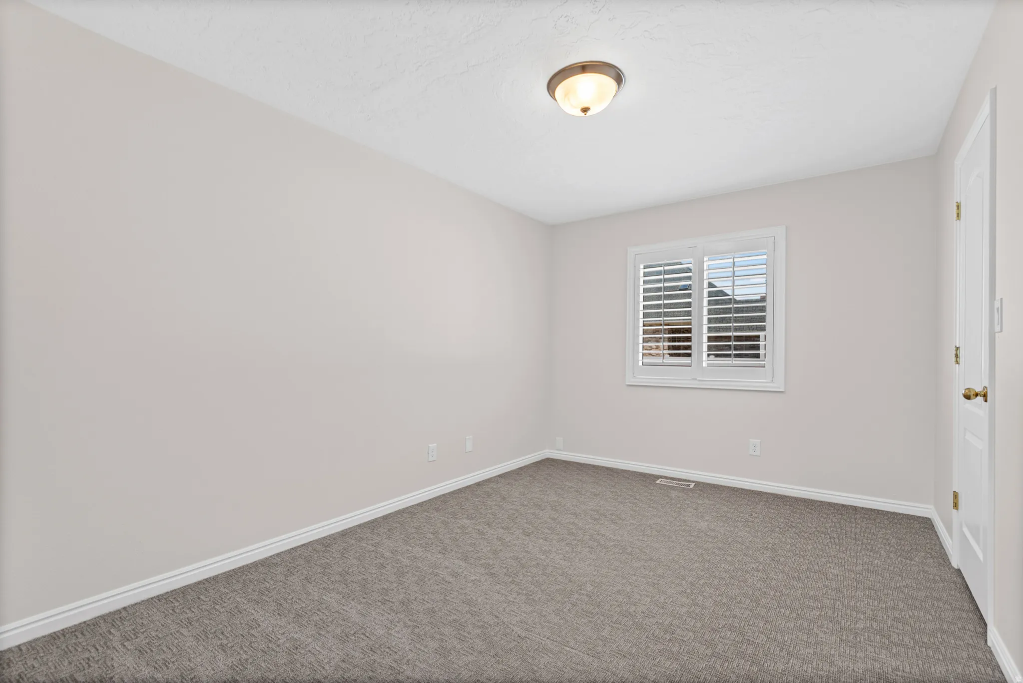 Neutral-toned room featuring a window with white plantation shutters, textured carpeting, and a flush-mount ceiling light fixture