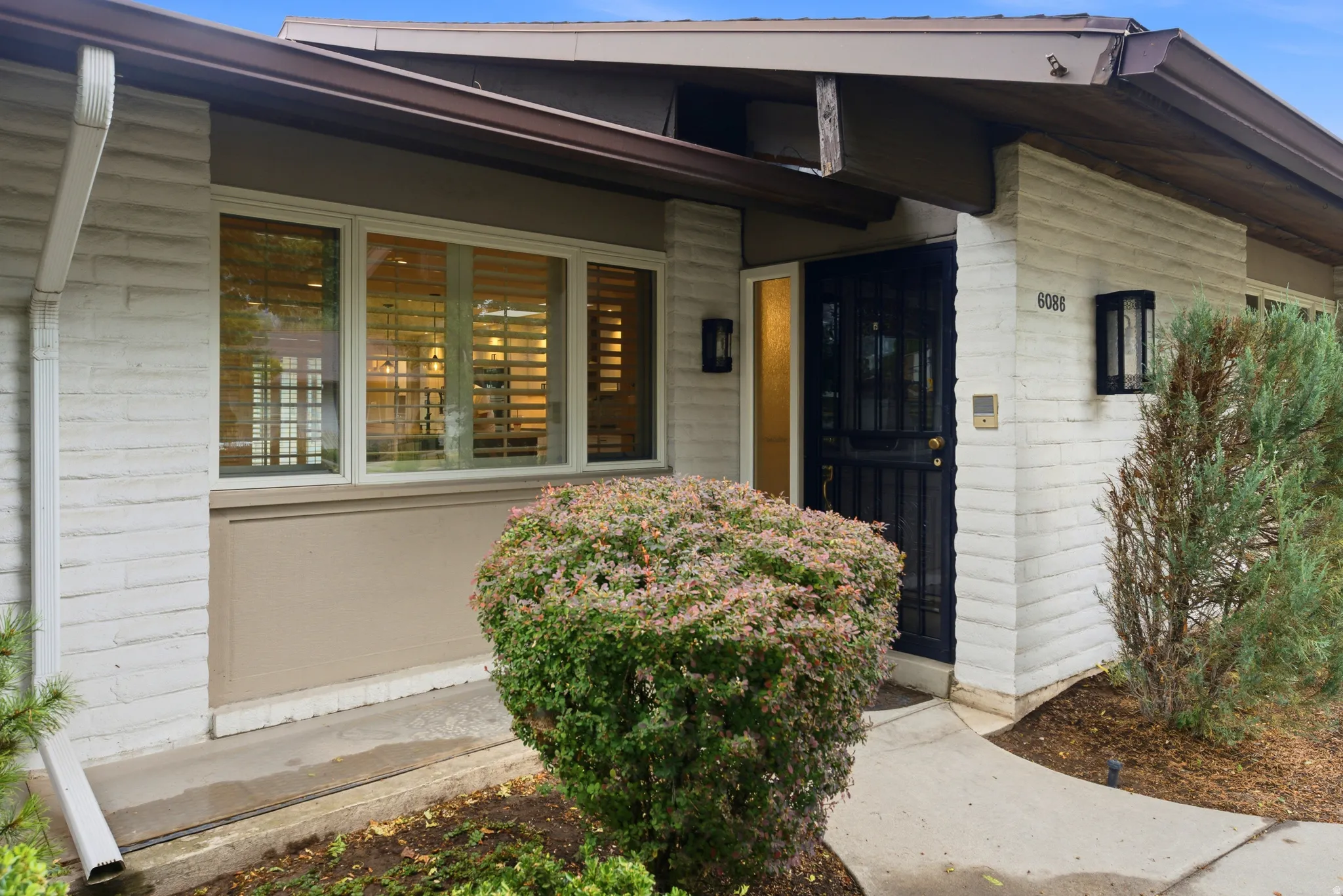 White brick exterior with a dark security door, featuring dual exterior wall sconces, address numbers, and a prominent front window with horizontal blinds