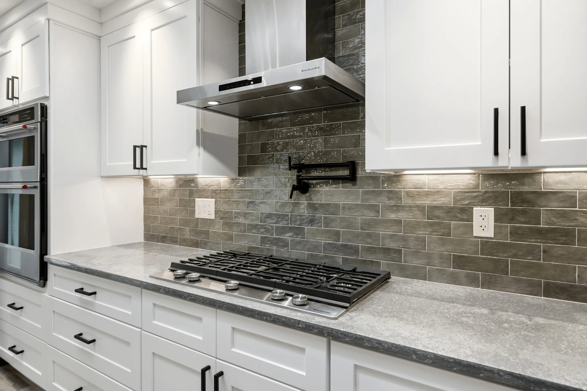 Modern kitchen featuring white shaker cabinetry with black hardware, a gas cooktop, stainless steel wall ovens, a coordinating range hood, and a grey subway tile backsplash