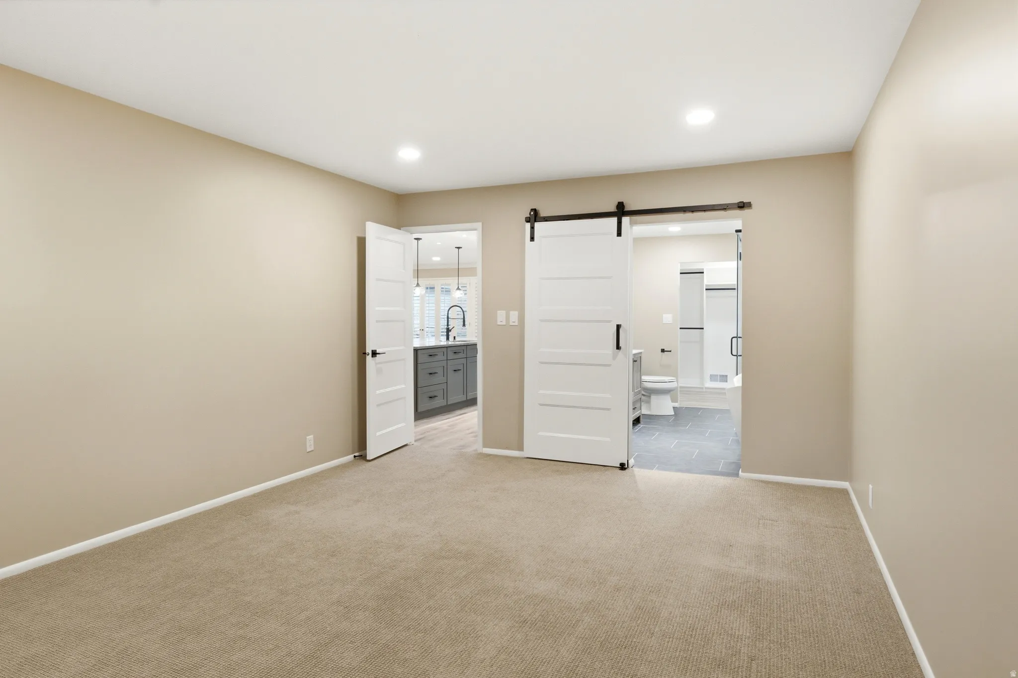 Spacious room featuring neutral carpeting, recessed lighting, and a modern barn door entry to a bathroom with tile flooring
