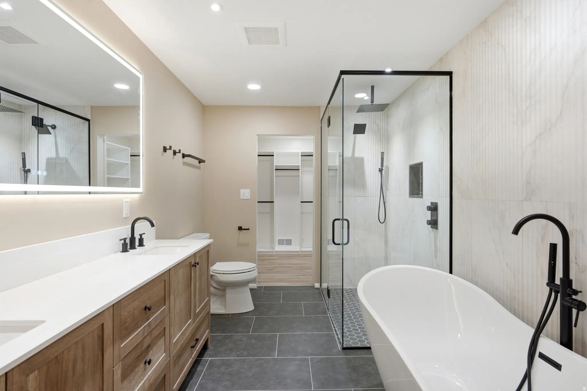 Modern bathroom featuring a freestanding soaking tub with a floor-mounted faucet, a glass-enclosed shower with a rain showerhead, a double vanity with a light wood-finish cabinet and white countertop, a backlit mirror, and dark tile flooring