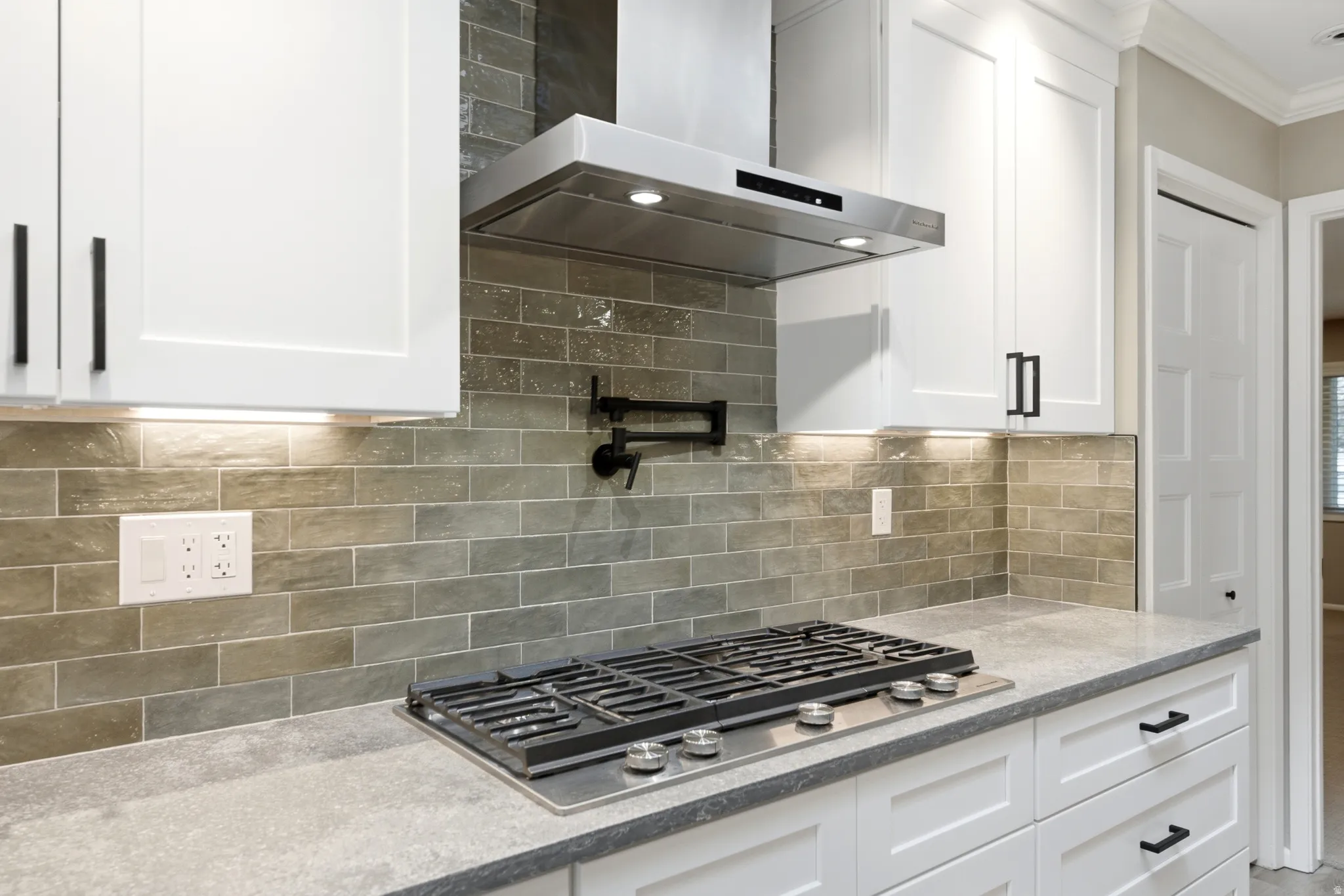 Modern kitchen featuring a gas range, stainless steel range hood, pot filler faucet, white shaker cabinetry with dark hardware, and a subway tile backsplash