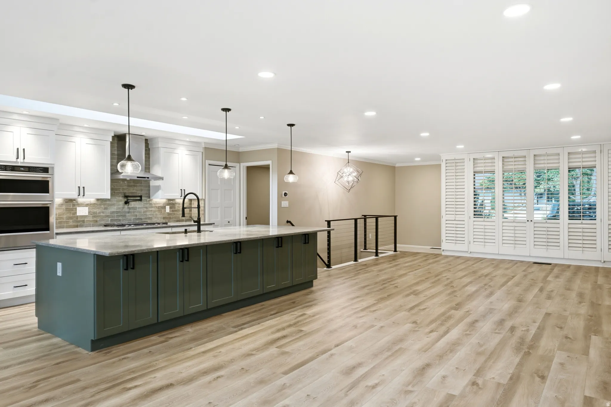 Spacious kitchen and living area featuring a large center island with a built-in sink, stainless steel appliances, light wood-finish flooring, recessed lighting, and white plantation shutters