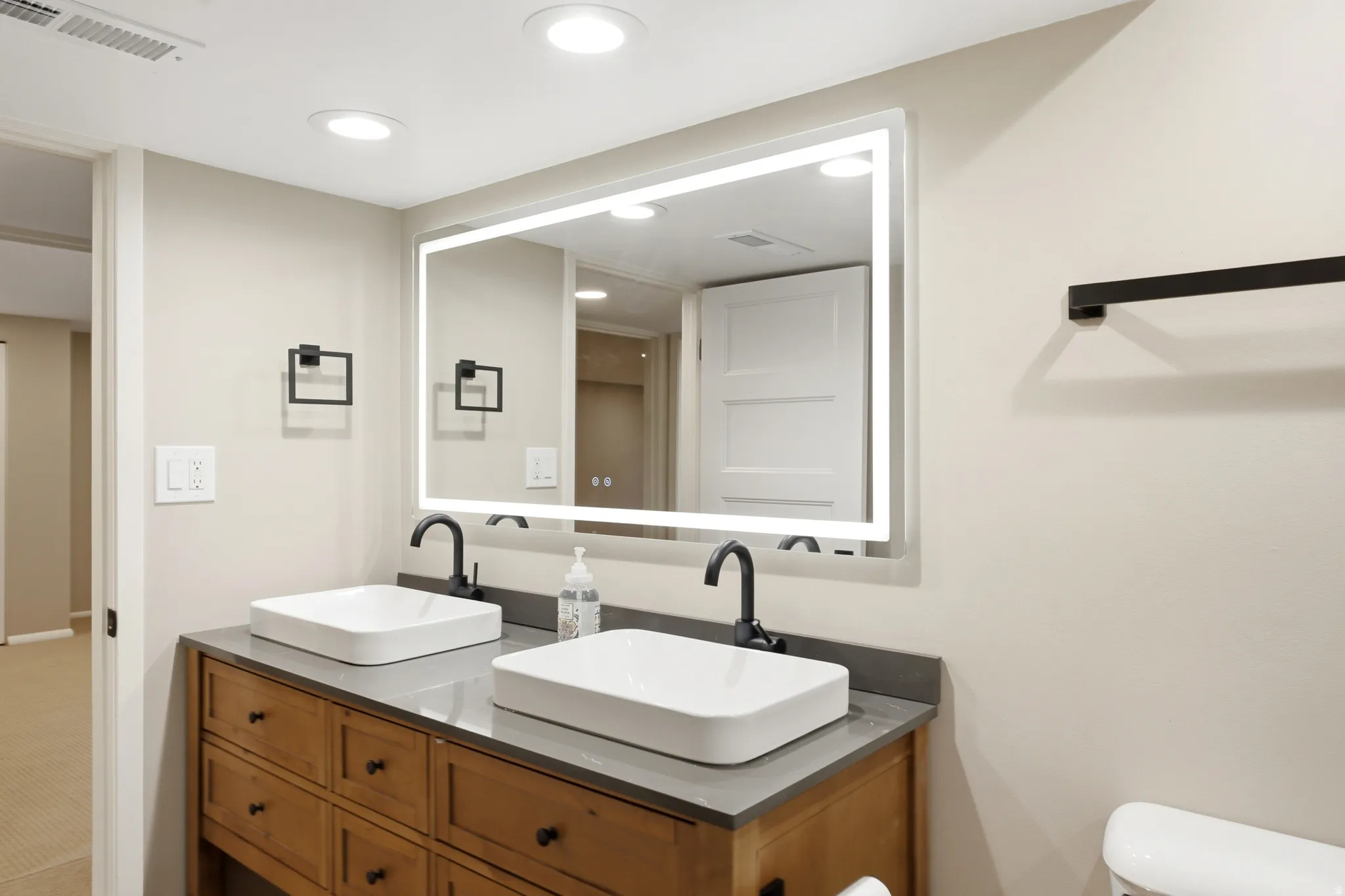 Dual vessel sinks with matte black fixtures, an illuminated mirror, and a wood-finish vanity with drawer storage