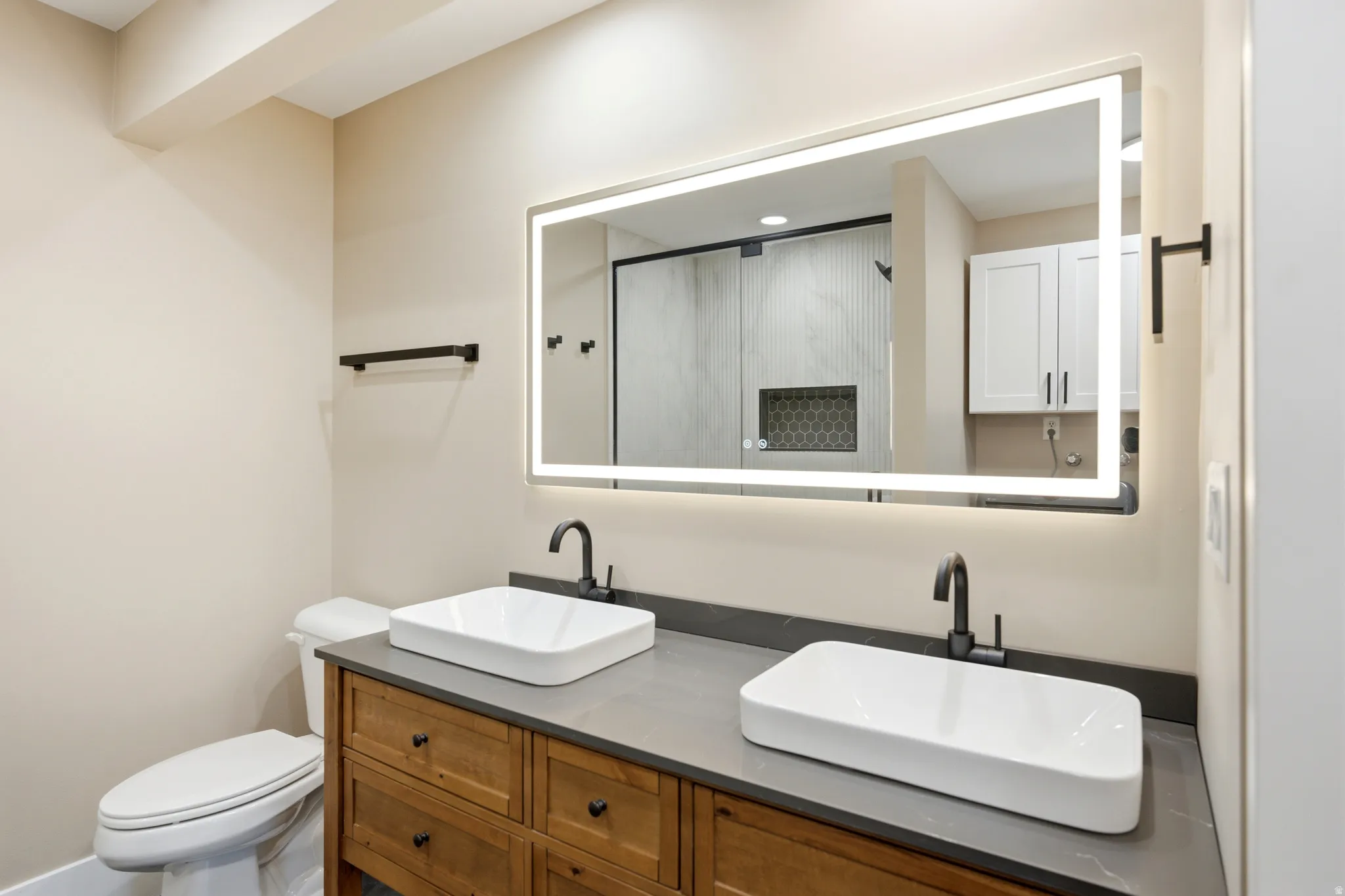 Dual vessel sinks set atop a wood-finish vanity with dark hardware, illuminated by a large backlit mirror