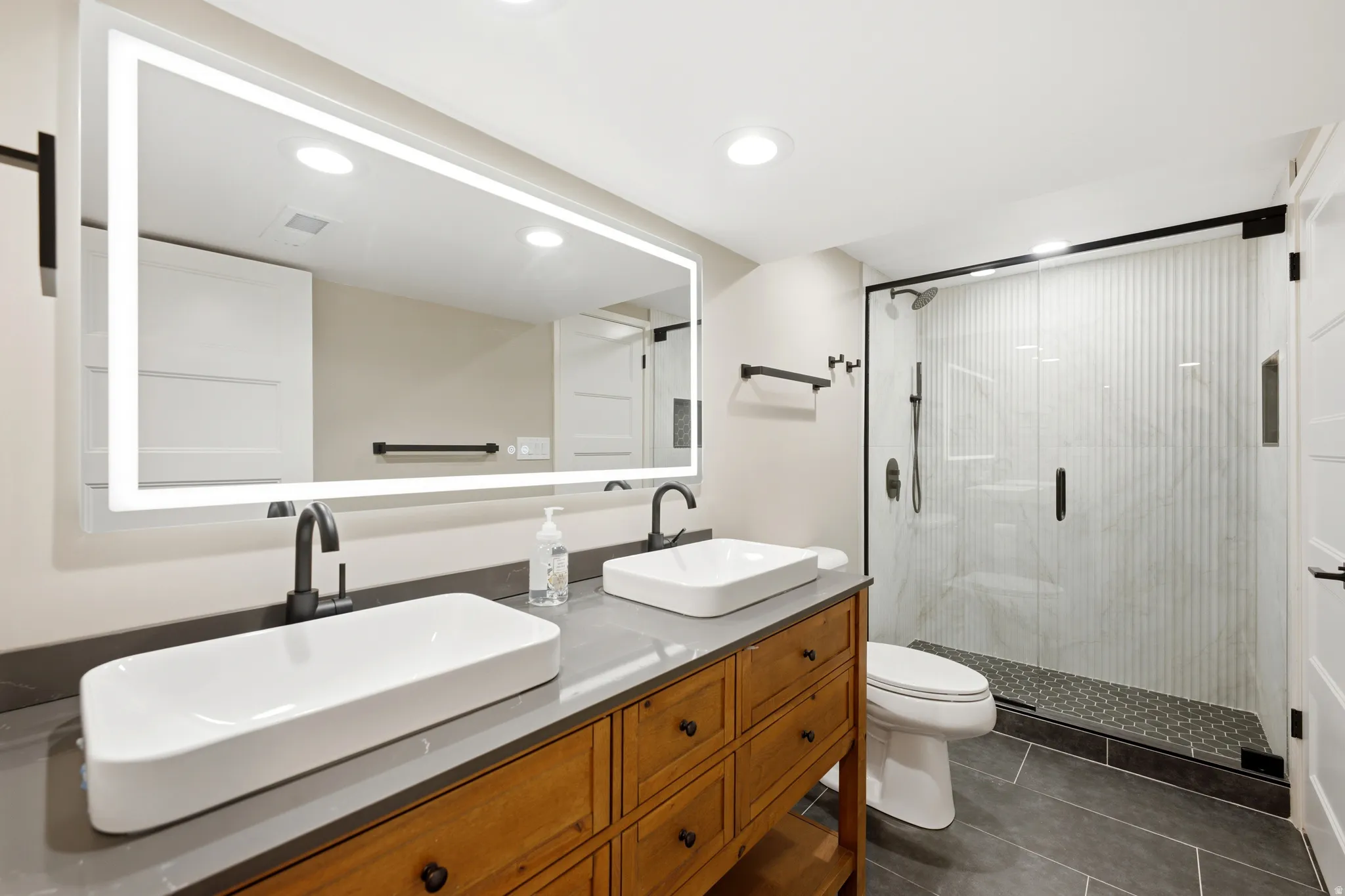 Bathroom featuring a wood-finish double vanity with rectangular vessel sinks, an illuminated mirror, a glass-enclosed shower with vertical accent tiling, and dark floor tiling