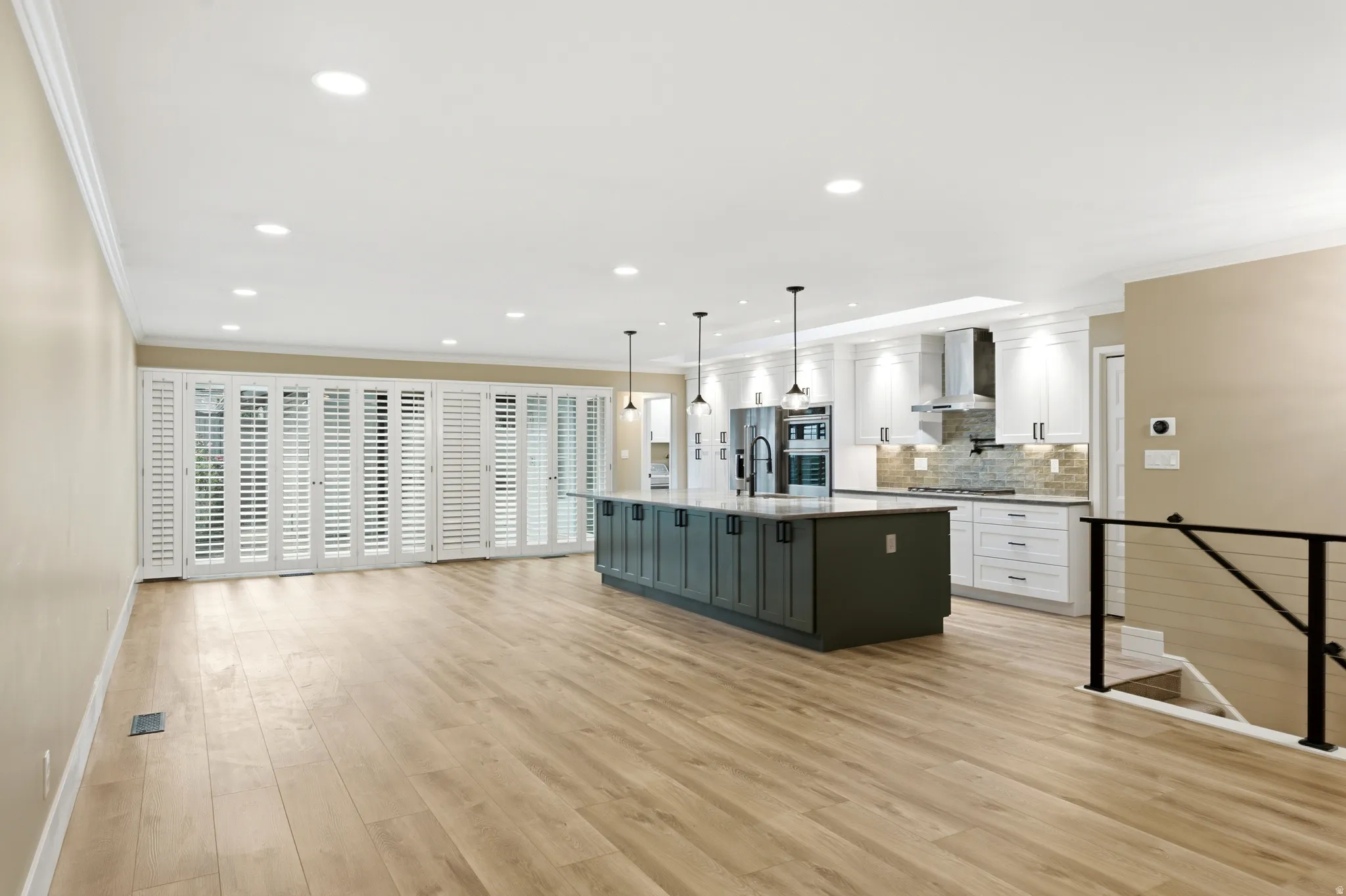 Spacious open-concept living area featuring wood-finish flooring, a contemporary kitchen with a dark-toned island and light cabinetry, stainless steel appliances, and a full wall of white plantation shutters