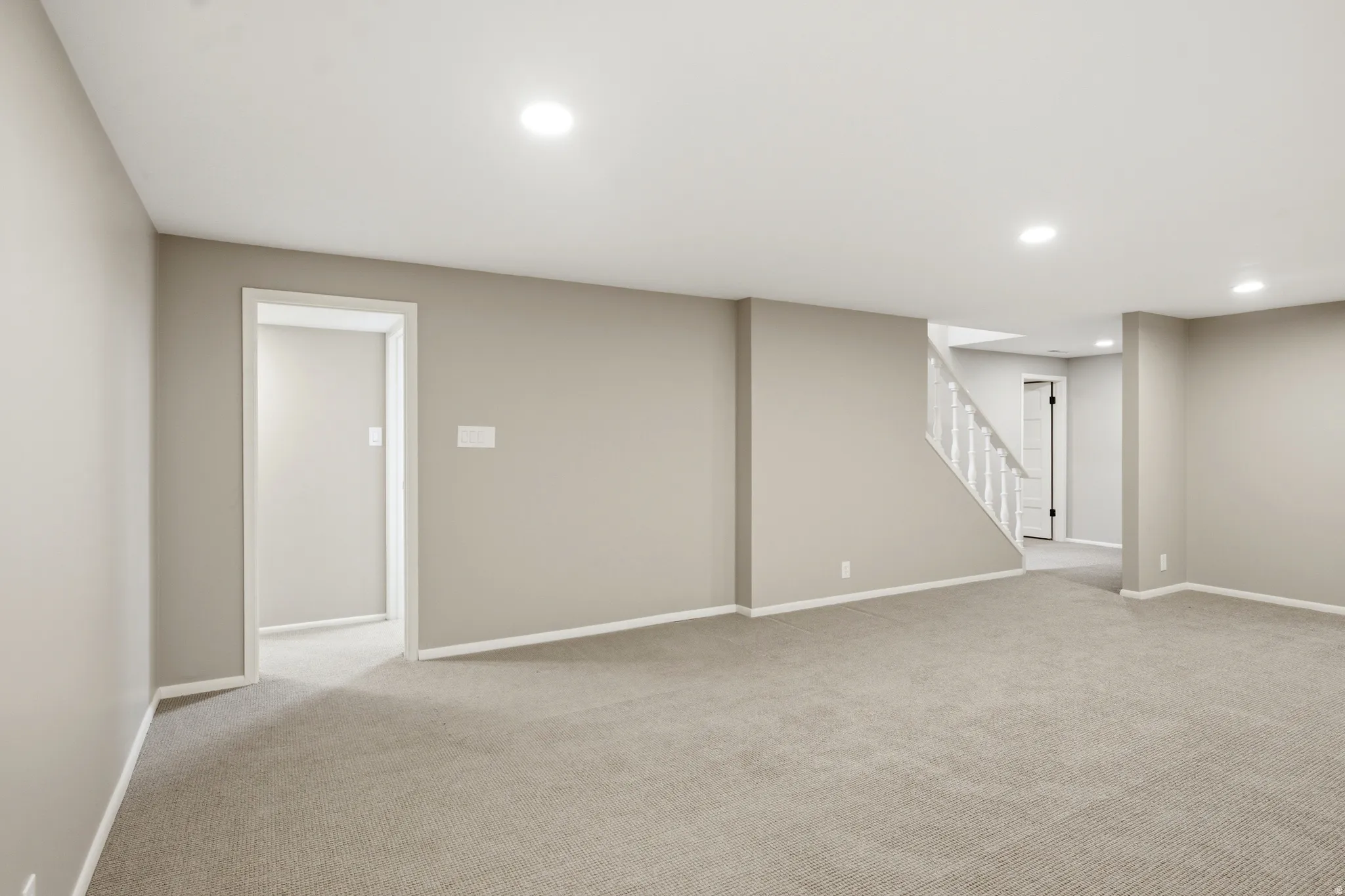 Spacious room featuring neutral carpeting, recessed lighting, light gray wall paint, white trim, and a white staircase with balusters
