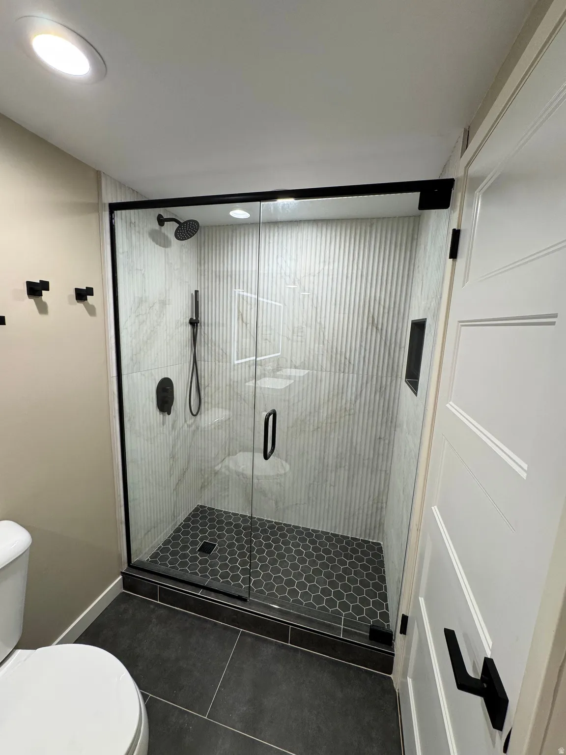 Contemporary shower enclosure featuring a frameless glass door with matte black hardware, hexagon tile flooring, and marble-look wall tiling with a textured vertical pattern