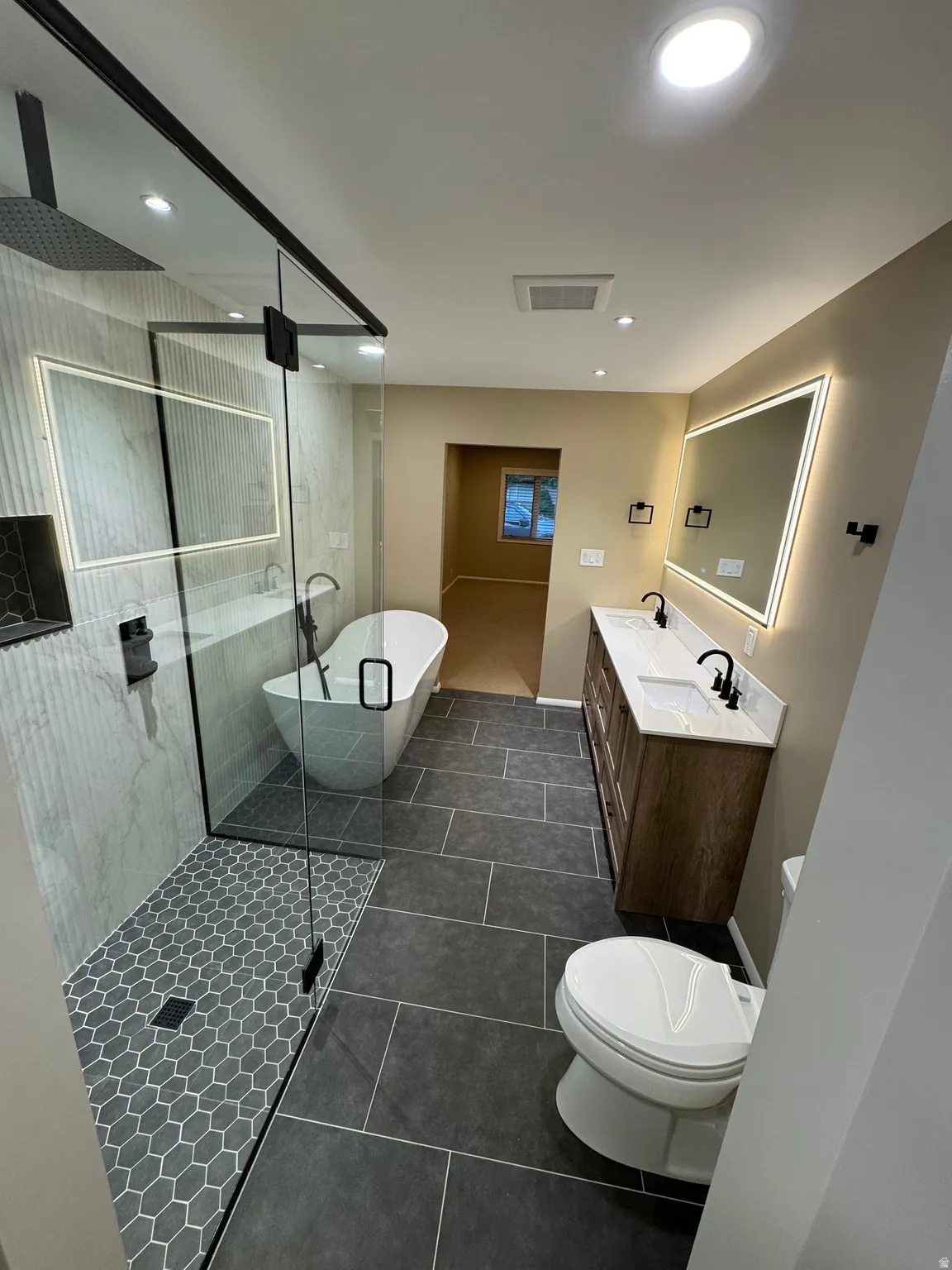 Contemporary bathroom featuring a spacious walk-in shower with a rainfall showerhead and hexagonal floor tiling, a freestanding soaking tub, a double vanity with stone countertops and undermount sinks, illuminated mirrors, and large format floor tiling