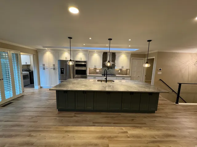 Modern kitchen featuring a large island with dark green cabinetry and a light-toned countertop, stainless steel appliances, white upper cabinetry, and wood-finish flooring