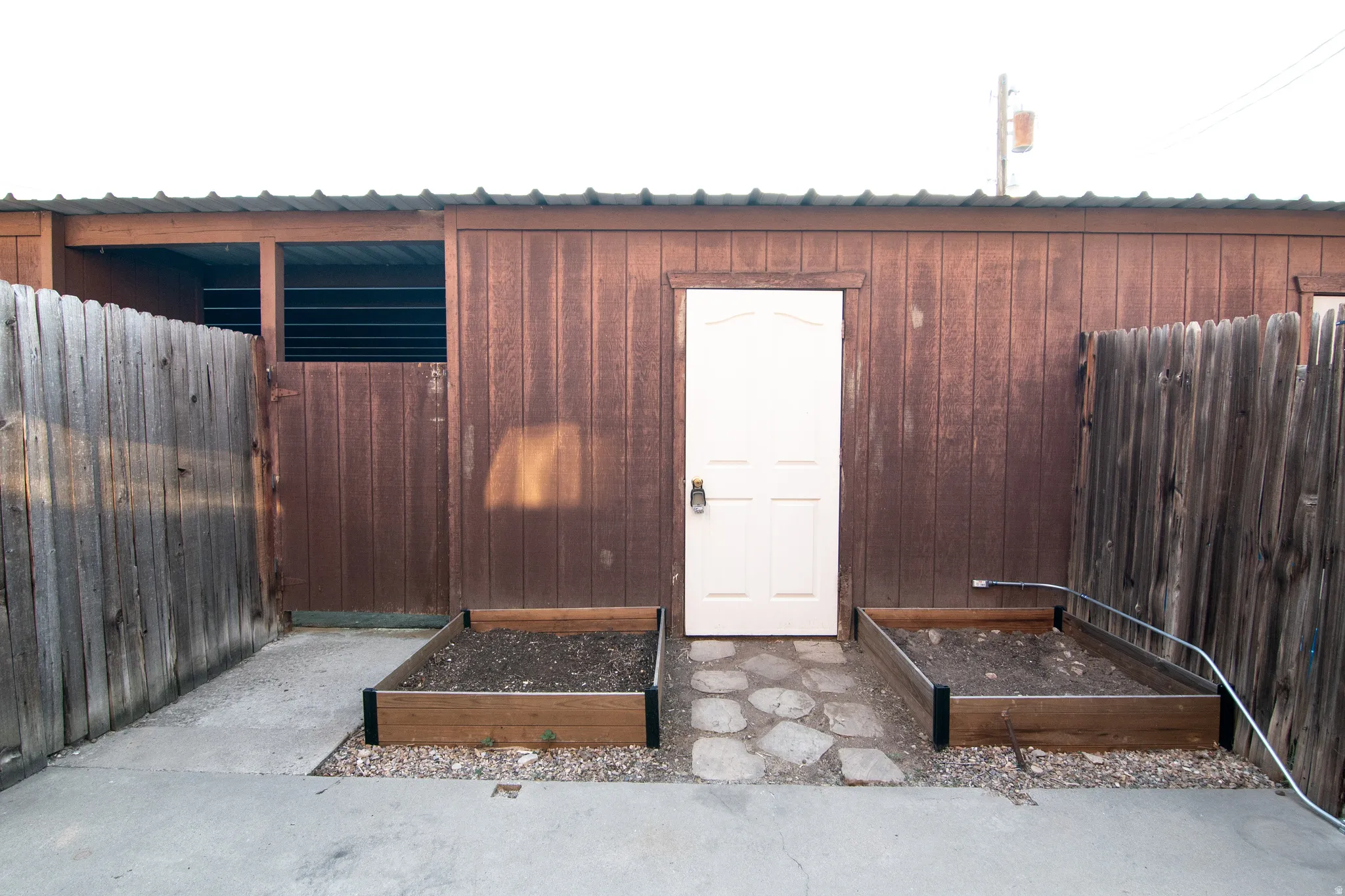 Wood-paneled outbuilding with a metal roof, white pedestrian door, and two raised garden beds