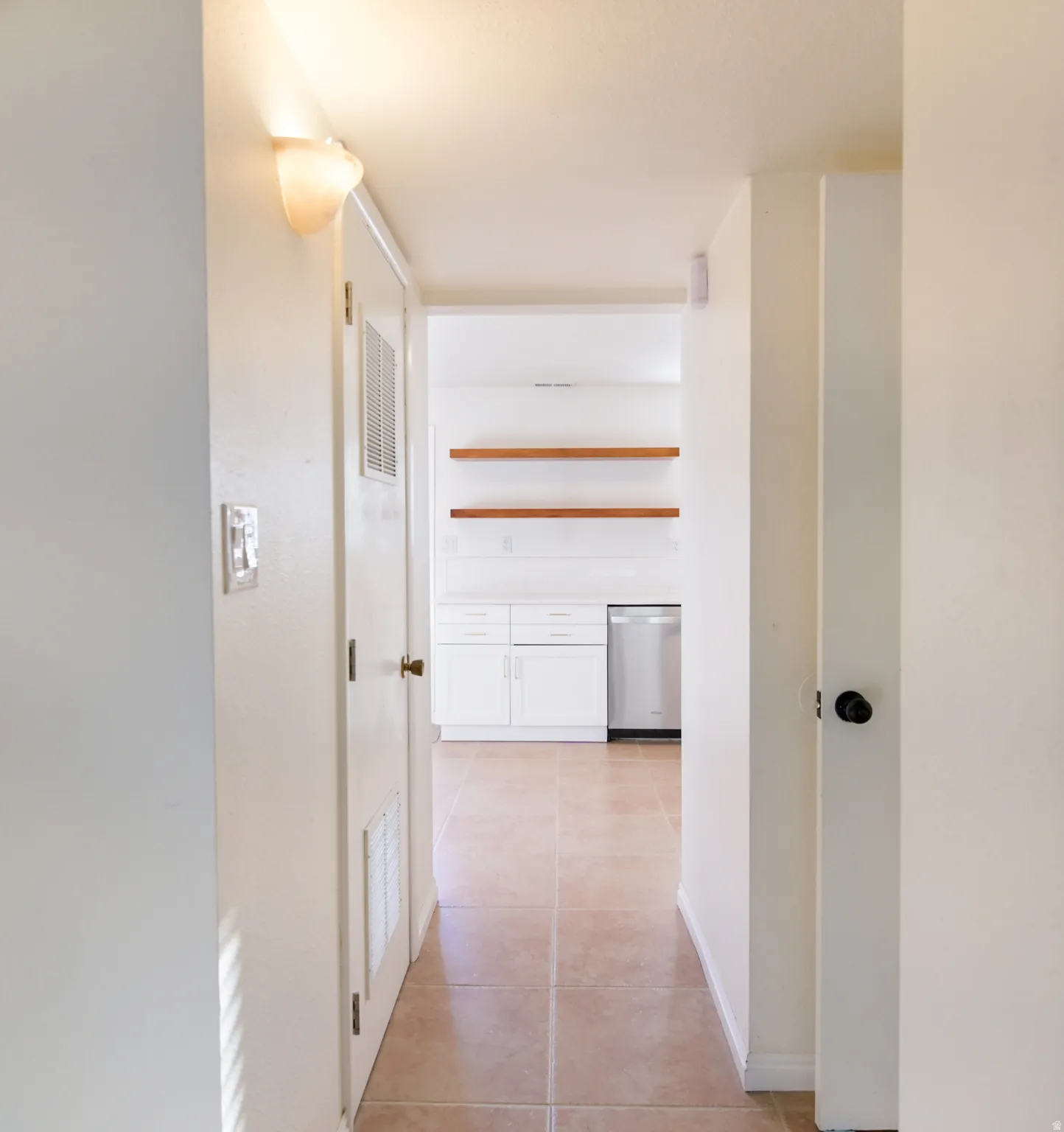 Interior hallway with tile flooring leading to a kitchen featuring white cabinetry, a stainless steel dishwasher, and two wood-finish floating shelves