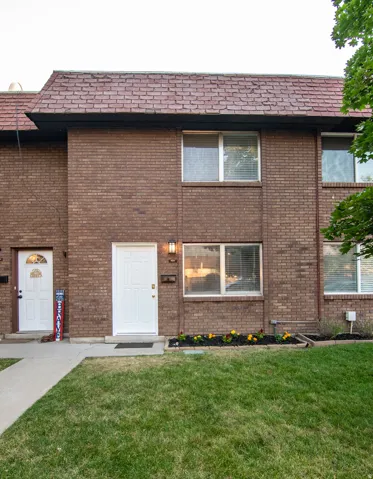 Brick exterior with a peaked roofline, featuring a white entrance door, a porch light fixture, and a mulched garden bed with flowers