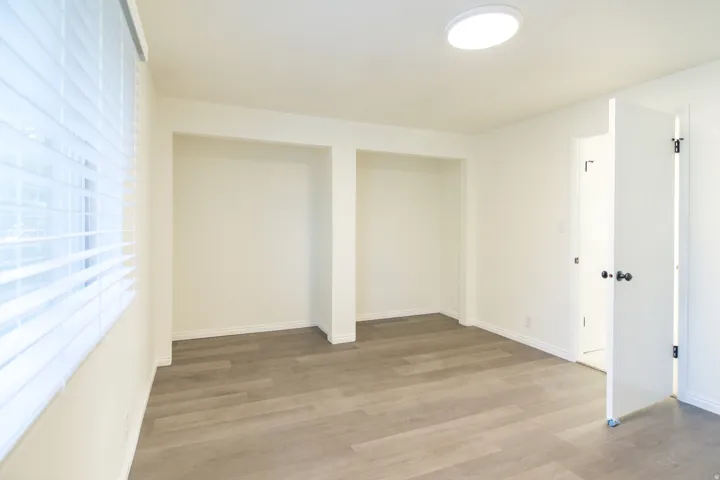 Bright room featuring wood-finish flooring, recessed wall alcoves, white window blinds, a flush-mount ceiling light, and a white door with black hardware