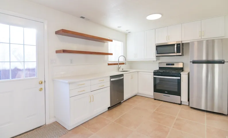 Kitchen featuring white cabinetry with gold-finish hardware, stainless steel appliances, a white subway tile backsplash, light-toned countertops, and wood-finish open shelving