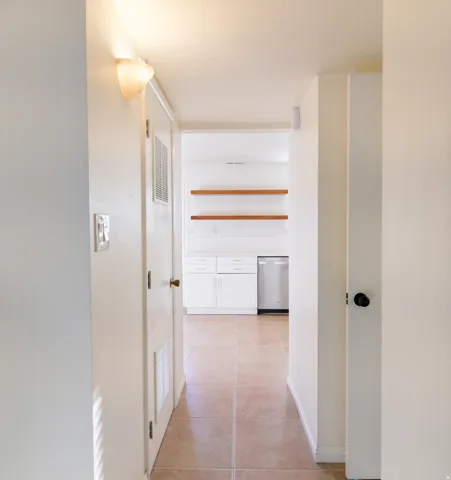 Interior hallway with tile flooring leading to a kitchen featuring white cabinetry, a stainless steel dishwasher, and two wood-finish floating shelves