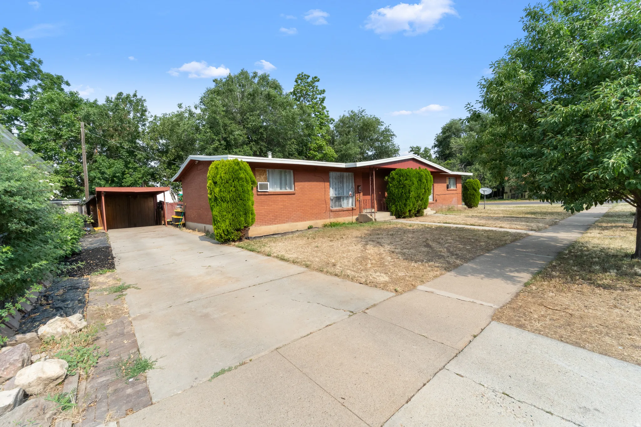 Brick exterior featuring a covered carport, mature trees, concrete driveway, and sidewalk access