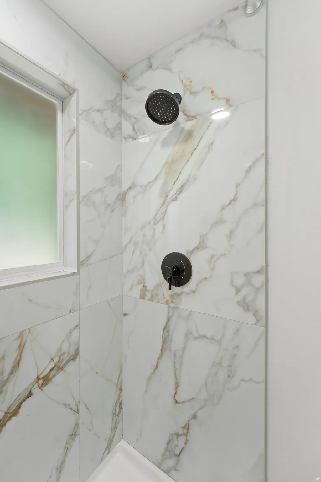 Shower enclosure featuring marble-look tile, matte black shower head, coordinating matte black control handle, and a frosted window with white trim