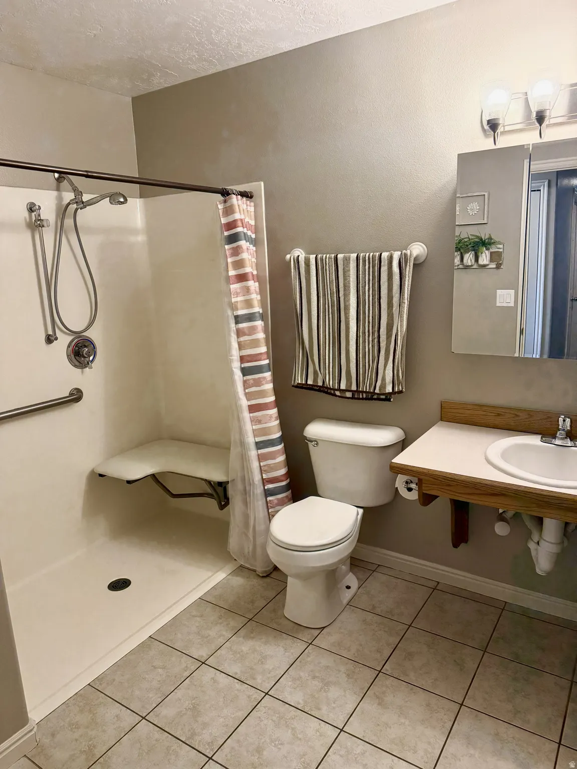Bathroom featuring a walk-in shower with a built-in seat and grab bar, tile flooring, a toilet, and a vanity with an integrated sink and wood-finish support