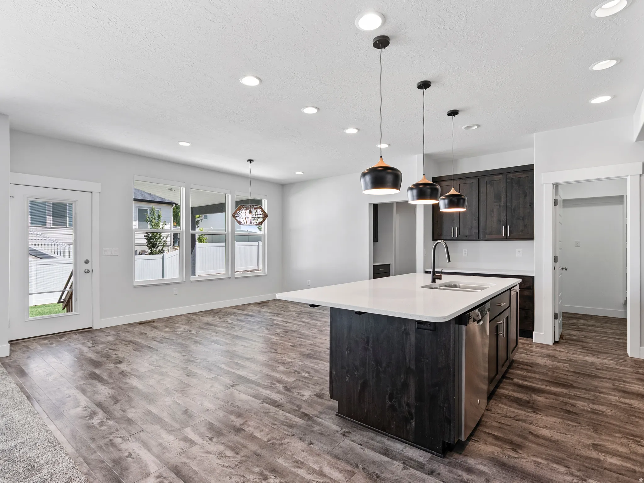 Kitchen island with a white countertop and integrated sink, stainless steel dishwasher, dark wood-finish cabinetry, recessed ceiling lights, and pendant lighting