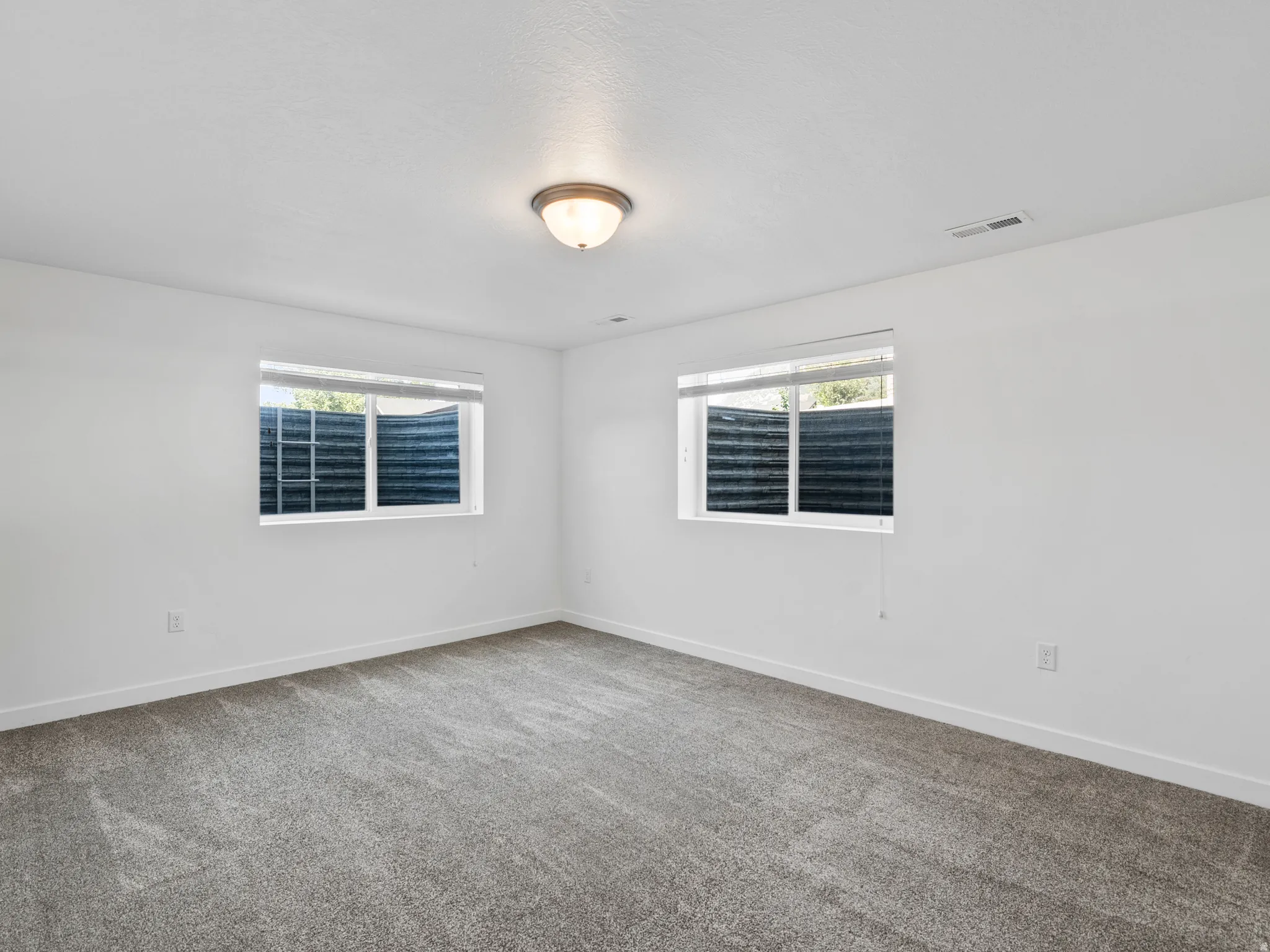 Carpeted room with white walls, two windows featuring horizontal blinds, and a flush-mount ceiling light fixture