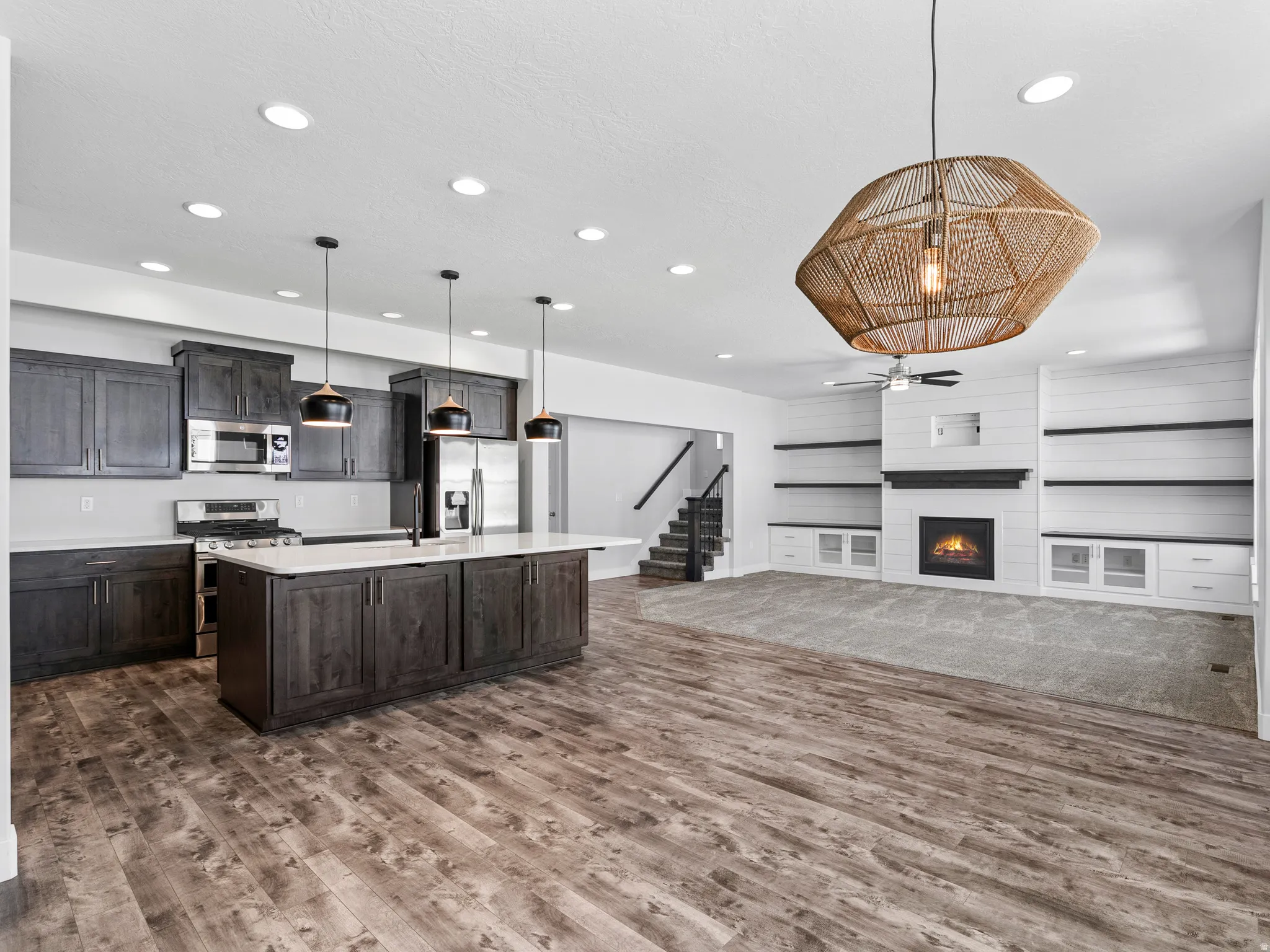 Open-concept living space featuring wood-finish flooring, a kitchen island with quartz countertops, dark wood cabinetry, a built-in fireplace with shiplap detailing, and integrated shelving with lower cabinetry
