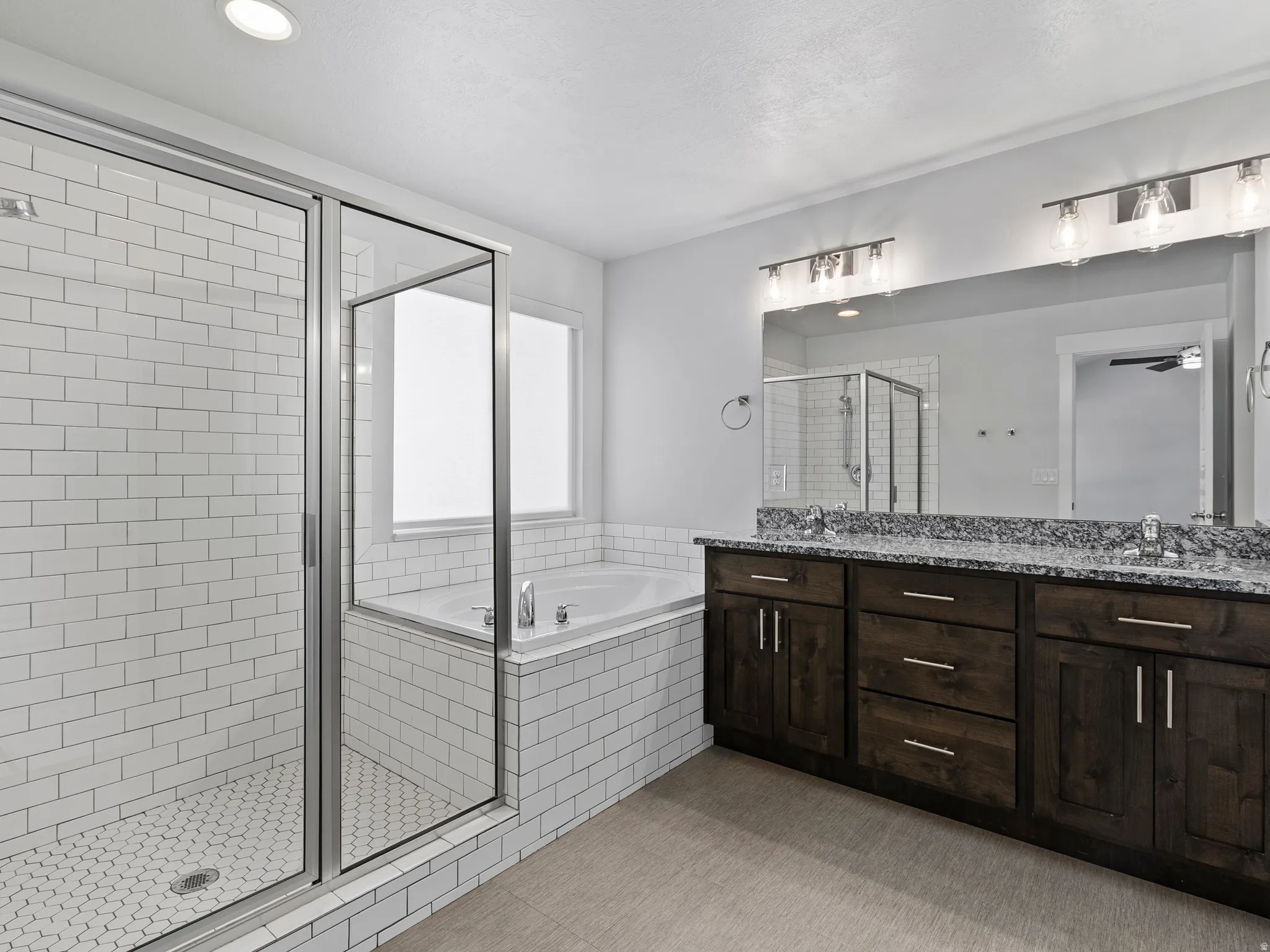 Spacious bathroom featuring a glass-enclosed shower with white subway tile, a built-in tub, dual sink vanity with a stone-finish countertop, dark wood-finish cabinetry, and recessed lighting