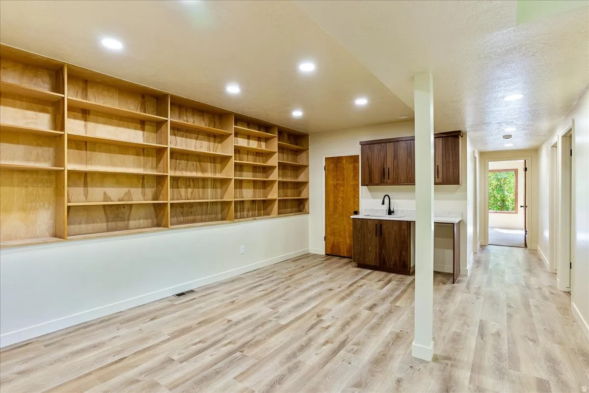Spacious room featuring extensive built-in wooden shelving, wood-finish flooring, recessed lighting, and a wet bar with dark wood cabinetry, a white countertop, and a matte black faucet
