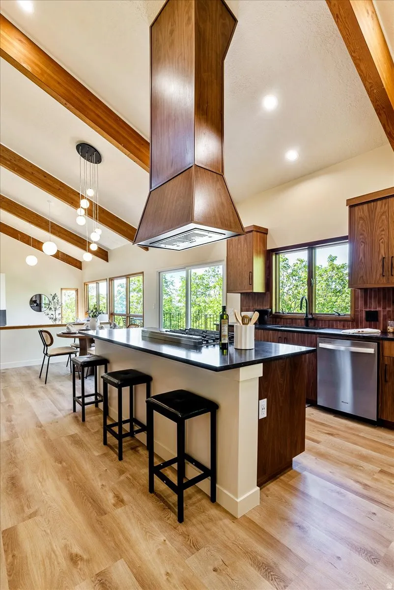 Open-concept kitchen featuring vaulted ceilings with exposed wood beams, a large island with dark countertops, wood-finish cabinetry, and stainless steel appliances
