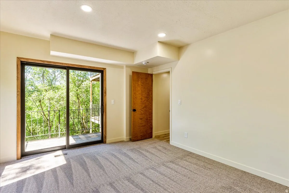 Carpeted room with a sliding glass door leading to a balcony, featuring a dark metal railing and surrounded by mature trees