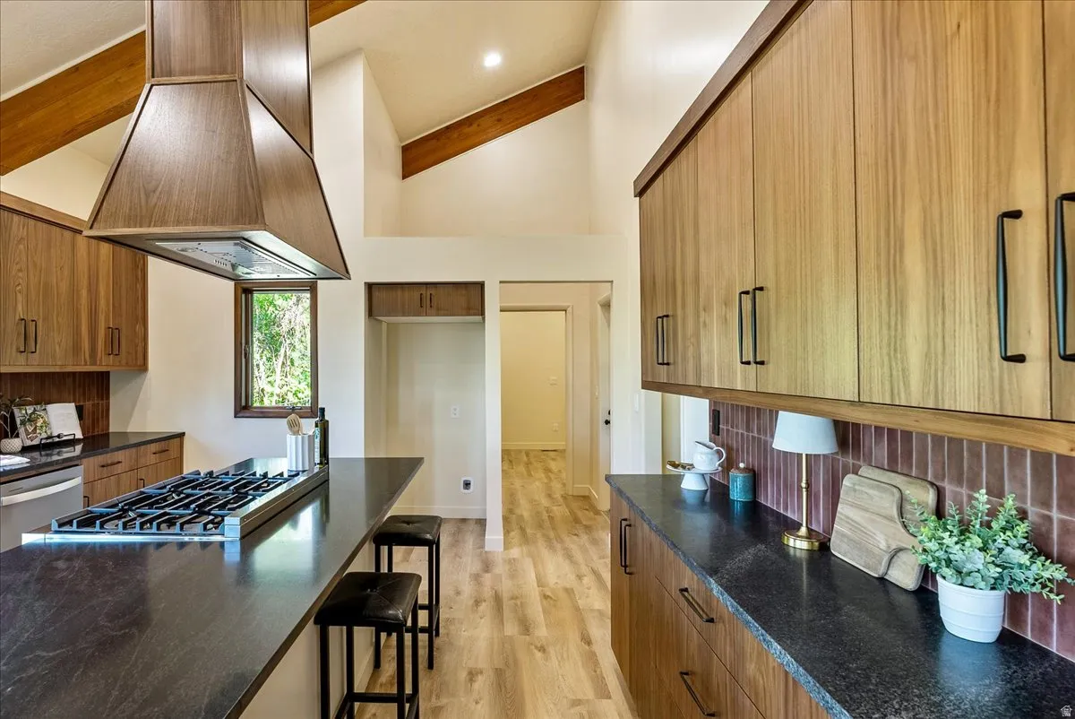 Spacious kitchen featuring a large island with a gas range, wood-finish cabinetry, dark countertops, a unique tile backsplash, and vaulted ceilings with exposed beams